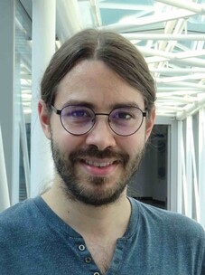 Louis Veyrat mit Brille und langen Haaren, vor einem modernen Architekturhintergrund.